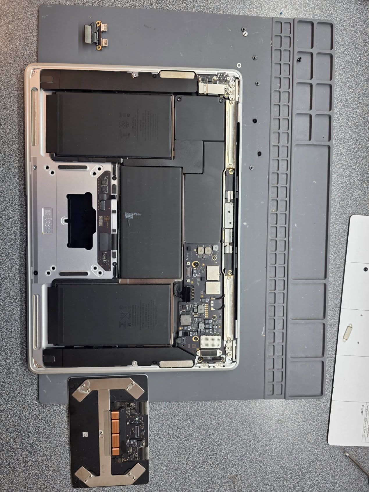 MacBook opened up on a Computer Wall iFixit project mat showing the motherboard, dual batteries, and removed trackpad — a System Exam in progress.